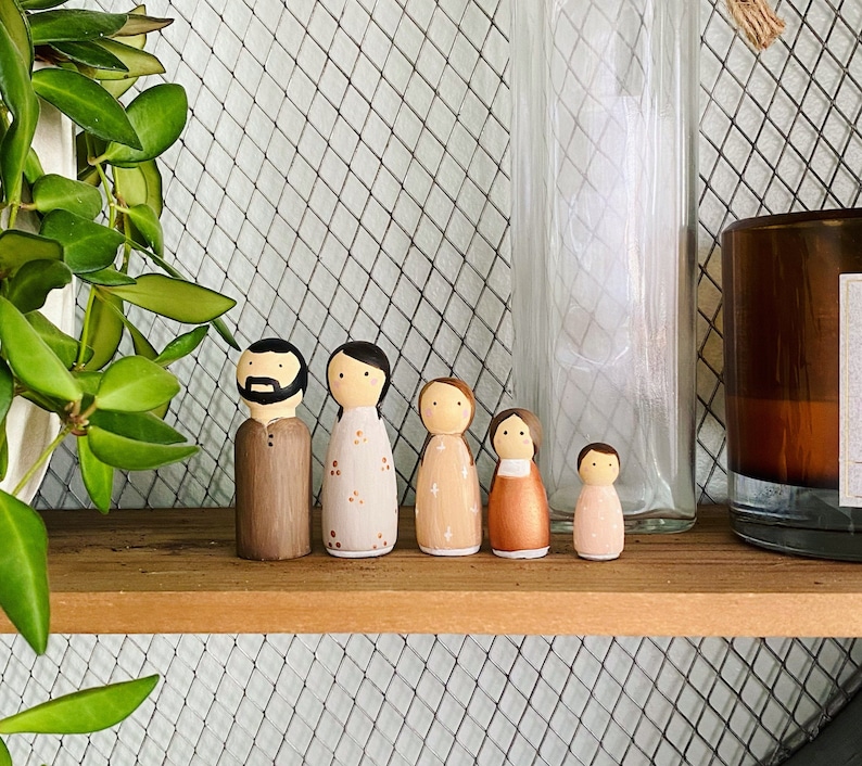 May include: A set of six wooden peg dolls, painted in various shades of brown, pink, and white. The dolls are standing on a wooden shelf with a wire mesh background.