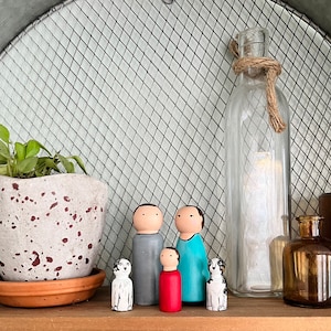 May include: A set of five wooden figurines, including two adults and a child, standing in front of a clear glass bottle with a rope tied around the neck. The figurines are painted in various colors, including gray, blue, and red. The child figurine is holding a red heart. The figurines are standing on a wooden surface.