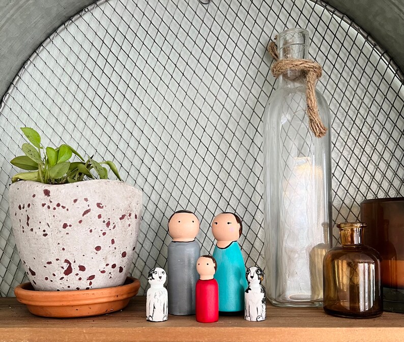 May include: A set of five wooden figurines, including two adults and a child, standing in front of a clear glass bottle with a rope tied around the neck. The figurines are painted in various colors, including gray, blue, and red. The child figurine is holding a red heart. The figurines are standing on a wooden surface.