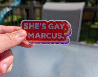 May include: A sticker with the text "SHE'S GAY, MARCUS." in white letters on a red background. The sticker has a rainbow-colored border and is held up against a blurred outdoor background.