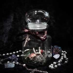 May include: A clear glass jar with a rounded lid, containing a miniature scene with moss, dried flowers, and a small bone. A brown ribbon and a silver cross adorn the jar. A rosary and crystals are placed around the jar.