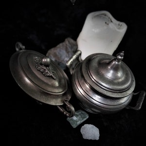 Peut inclure: Deux récipients antiques argentés avec couvercles, poignées ornées et un motif de feuille sur le couvercle, sur fond sombre. Petites pierres et un objet blanc de forme irrégulière sont également présents.