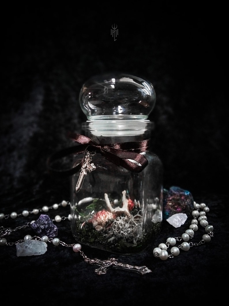 May include: A clear glass jar with a rounded lid, containing a miniature scene with moss, dried flowers, and a small bone. A brown ribbon and a silver cross adorn the jar. A rosary and crystals are placed around the jar.