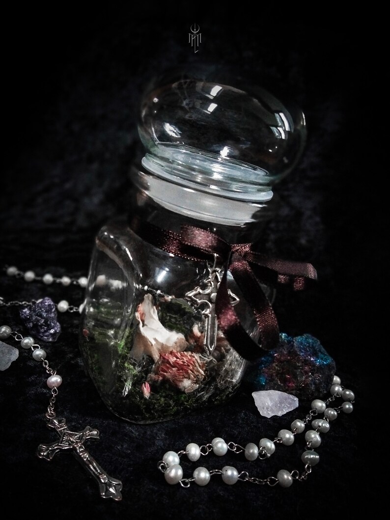 May include: A clear glass jar with a rounded lid, filled with dried flowers and moss, tied with a brown ribbon. A silver cross charm hangs from the ribbon. A pearl rosary and crystals are arranged around the jar on a dark surface.