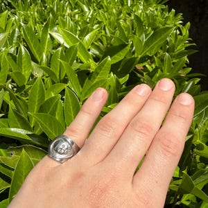 May include: A silver-toned metal ring featuring a raised, circular relief design of a human face on the front surface. The ring has a smooth, polished band that tapers slightly toward the back, showcasing a metallic finish and a distinct sculptural detail.