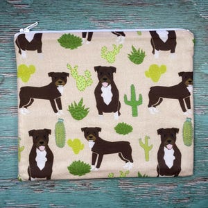 May include: A beige zippered pouch featuring a repeating pattern of brown dogs with white markings, green cacti, and other desert plants. The pouch has a white zipper and is likely made of fabric. The design is playful and suitable for pet lovers.