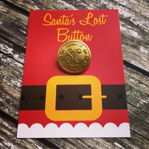 May include: A festive Christmas card with a red backdrop, showcasing a gold-coloured button inscribed with "Property of SC North Pole." The card design includes a brown belt, a yellow buckle, and the text "Santa's Lost Button" in yellow.