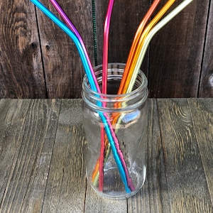 May include: A glass jar filled with six colorful metal drinking straws. The straws are bent and are in various shades of blue, pink, orange, and yellow.
