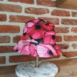 Op de afbeelding: Een roze bucket hat met een bloemenpatroon van rode en roze bloemen. De hoed wordt gepresenteerd op een marmeren standaard met een goudkleurige metalen staaf, tegen een bakstenen muur en een houten plank.
