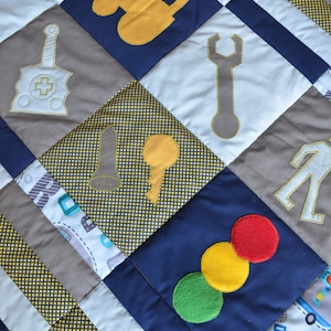 May include: A patchwork quilt with a blue, white, and gray color scheme. The quilt features various embroidered images of tools, including a wrench, a key, a traffic light, and a tractor. The quilt is made of different fabrics with a yellow and black polka dot pattern.