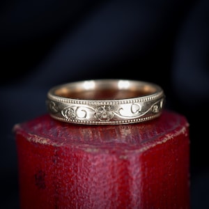 Puede incluir: Un anillo dorado con un diseño floral y de vid. El anillo presenta una serie de pequeños puntos en relieve a lo largo de los bordes superior e inferior. El diseño floral incluye flores detalladas y vides onduladas. El anillo se encuentra sobre un fondo oscuro.