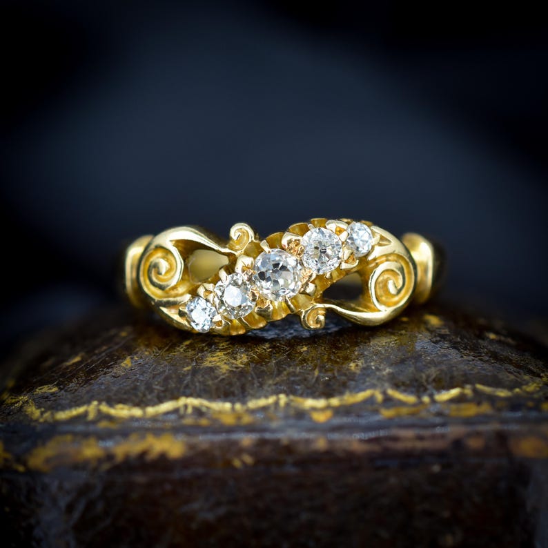May include: A gold ring with five clear gemstones set in a decorative, swirling design. The ring has a vintage aesthetic, with ornate detailing and a polished finish. The gemstones are round-cut and sparkle against the gold setting. A piece of jewelry.