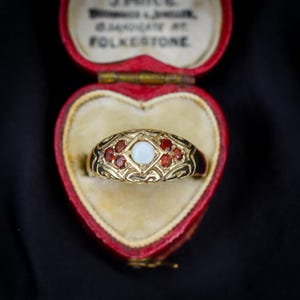 Può includere: Un anello d'oro con una gemma bianca centrale e gemme rosse circostanti, incastonato in un design ornato. L'anello è presentato in un cofanetto di velluto rosso a forma di cuore. Il design dell'anello include intricati motivi a filigrana.