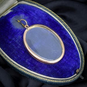 May include: A gold-toned oval pendant with a clear glass center, set in a gold frame. The pendant is displayed in a blue velvet jewelry box with a gold trim. The pendant has a small loop at the top for hanging.