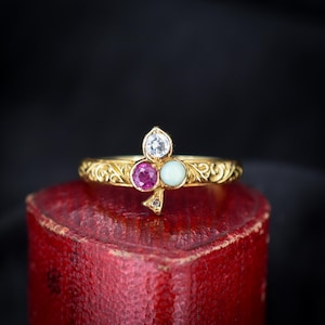 May include: A gold ring with an ornate band and a cluster of three gemstones: a clear, teardrop-shaped stone, a round pink stone, and a round light blue stone. The ring is set on a red surface.