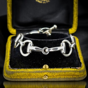 May include: A polished sterling silver chain bracelet featuring repeating horse bit links. The bracelet is constructed with a toggle clasp mechanism and displays a smooth, reflective metallic finish throughout the entire jewelry piece.