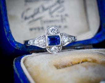 Può includere: Un anello color argento con uno zaffiro blu rettangolare centrale affiancato da due diamanti rotondi sopra e sotto, con ulteriori piccoli diamanti incastonati lungo le spalle affusolate della fascia, presentato in un portagioie in velluto blu.