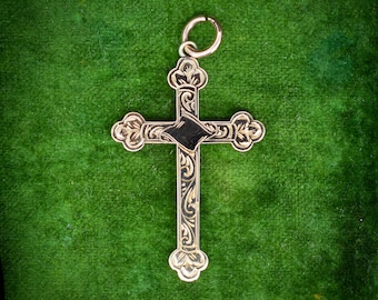 May include: A gold-coloured cross pendant with detailed floral and scroll patterns. The cross has a dark, diamond-shaped centre and a small loop for a chain. The pendant is set against a green backdrop, highlighting its ornate design.