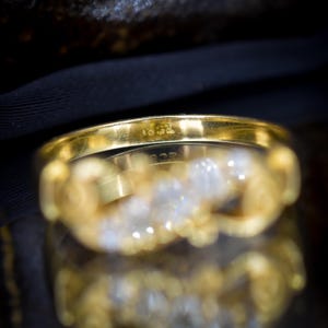 May include: Close-up of a gold ring with a row of clear gemstones. The ring has a smooth, polished band with markings. The gemstones are set in a decorative, ornate setting. The ring is displayed against a dark background.