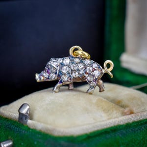 May include: A pig-shaped pendant crafted from silver and gold, featuring a body encrusted with numerous rose-cut diamonds and small red gemstone accents. The charm includes a small gold jump ring at the top for attachment to a chain or bracelet.