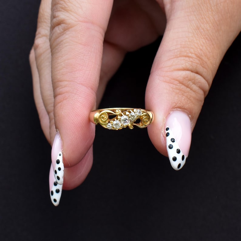 May include: A gold ring with a decorative filigree design and several small, clear gemstones. The ring is held between fingers against a black background. The ring's design includes swirling patterns.