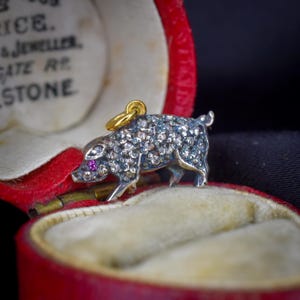 May include: A silver pig charm featuring a textured body with numerous small, sparkling stones and a single vibrant pink stone for the eye. The charm includes a small gold-toned metal loop at the top for attachment, presented inside a red jewellery box.