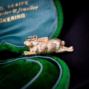 Könnte beinhalten: Ein goldfarbener Metall-Hasenanhänger mit detaillierter Fellstruktur und einem kleinen runden Biegering an der Oberseite. Der Hase ist in einer horizontalen, springenden Haltung dargestellt, mit nach vorne gestreckten Vorderpfoten und ausgestreckten Hinterbeinen.