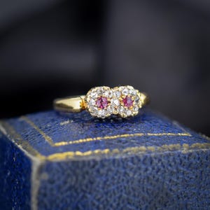 May include: A gold ring featuring two floral-shaped clusters, each centered with a round pink gemstone surrounded by small white stones. The band is smooth and polished gold, showcasing a detailed setting that holds the two central floral motifs.