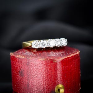 May include: A gold band ring featuring five round brilliant cut stones set in a row. The ring is positioned on top of a textured red cylindrical object. The stones are clear and reflective, catching the light against the dark background.