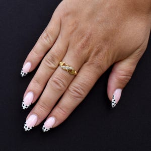 May include: A gold ring with a decorative design and small clear stones. The ring is on a person's hand with a black background. The nails are painted with a pink and white design with black polka dots.