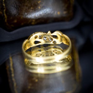 May include: Close-up of a gold ring with several small diamonds set into the band. The ring has an ornate design with a series of cut-out shapes. The ring is sitting on a dark surface.