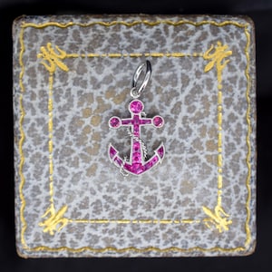 May include: A silver anchor pendant with pink gemstones, presented on a textured grey box with gold detailing. The anchor design features a rope and is set against a dark background.