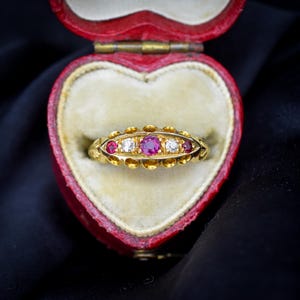 May include: A gold ring featuring a central round-cut pink gemstone flanked by two clear round-cut stones and two smaller pink round-cut stones, all set in a decorative gold band with scalloped edges, presented inside a red heart-shaped box.
