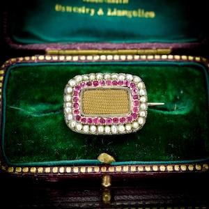 Pode incluir: Broche antigo com um centro retangular dourado, rodeado por uma fila de pedras preciosas rosa e uma borda de pequenas pérolas. O broche está engastado numa moldura prateada e apresentado numa caixa de joias de veludo verde.