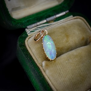 May include: An oval-shaped cabochon opal pendant set in a yellow gold prong setting. The opal displays a play of color with flashes of green and blue. A small circular gold jump ring is attached to the top of the pendant for hanging purposes.