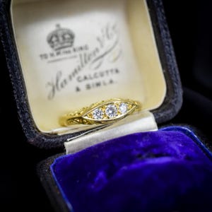 May include: A gold ring featuring three round clear stones set in a marquise-shaped frame, resting inside a dark blue velvet-lined jewelry box with the text TO H.M. THE KING Hamilton & Co CALCUTTA & SIMLA printed on the cream-colored interior lid.