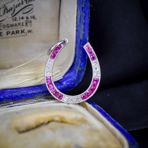 May include: A horseshoe-shaped pendant crafted from silver-toned metal, featuring alternating channel-set square-cut vibrant pink gemstones and round brilliant-cut clear white stones, finished with a small circular bail at the top for suspension.