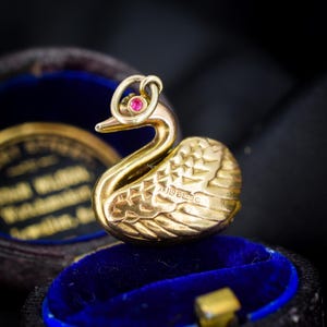 May include: A gold-toned swan charm featuring a small, circular pink gemstone set within the eye area. The swan has detailed, textured wing feathers and a small metal loop at the top for attachment. Hallmarks are visible on the side of the body.