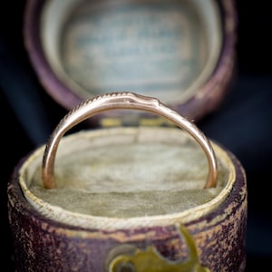 Puede incluir: Un anillo de oro con un diseño delicado y grabado, presentado en un joyero morado vintage con interior crema. La banda del anillo presenta una serie de pequeñas marcas espaciadas uniformemente. El joyero tiene un cierre de latón.