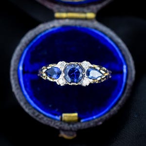 Könnte beinhalten: Ein Ring mit einem zentralen, rund geschliffenen blauen Saphir, flankiert von zwei tropfenförmig geschliffenen blauen Saphiren und vier kleinen, rund geschliffenen Diamanten in einer Ringschiene aus Gelbgold mit texturiertem Muster.