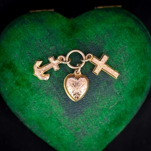 Może przedstawiać: Złoty naszyjnik z zawieszką w kształcie serca, kotwicy i dwoma krzyżami. Naszyjnik jest prezentowany w zielonym, aksamitnym pudełku w kształcie serca.