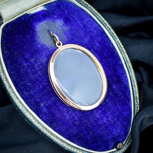 May include: A gold-toned oval pendant with a gray center, displayed in a blue velvet jewelry box. The pendant has a simple, elegant design with a polished finish. The box has a decorative trim.