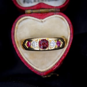 May include: A gold ring with a central square-cut red gemstone, flanked by two round white gemstones and two smaller red gemstones. The ring is set in a gold band and displayed in a red heart-shaped box.