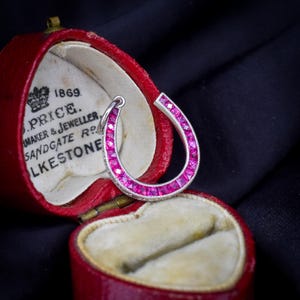 May include: A horseshoe-shaped pendant featuring a channel setting of vibrant pink square-cut stones framed in silver metal. The pendant rests inside a red heart-shaped jewelry box with an interior lining printed with the text 1869 J. PRICE.