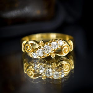 May include: A gold ring with five clear gemstones set in a decorative, swirling design. The ring is highly reflective, with its mirrored image visible below. The ring's band is also gold, and the overall aesthetic is ornate and elegant.