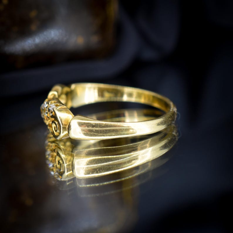 May include: A close-up of a gold ring with a decorative design. The ring has a band of gold and features small, sparkling stones. The ring is displayed on a reflective surface, with its reflection visible below.