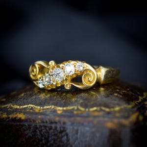 May include: A gold ring with five clear diamonds set in a decorative, swirling design. The ring is set on a dark, textured surface, with a dark background. The ring's band is a simple, polished gold.