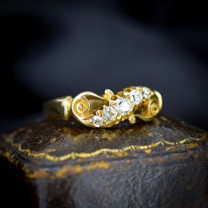 May include: A gold ring with a series of small diamonds set in a decorative, swirling design. The ring has a polished finish and is set against a dark background, highlighting the sparkle of the gemstones. The ring is likely an antique or vintage piece.