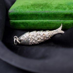 Könnte beinhalten: Ein silberner Fisch-Anhänger mit beweglichen Segmenten und detaillierten Schuppengravuren. Der Fisch hat ein offenes Maul mit einem kleinen, runden Biegering zum Aufhängen. Die metallische Oberfläche reflektiert das Licht und betont das Muster.