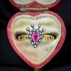 Könnte beinhalten: Ein Vintage-Ring mit einem herzförmigen Rubin als Mittelstein, umgeben von kleinen Diamanten und gefasst in einem Silber- und Goldband. Der Ring wird in einer roten herzförmigen Schmuckschatulle mit cremefarbenem Innenraum präsentiert.